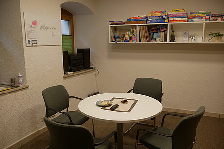 Raum mit einem Tisch und vier Stühlen, im Hintergrund befindet sich ein Wandregal mit Spielen.