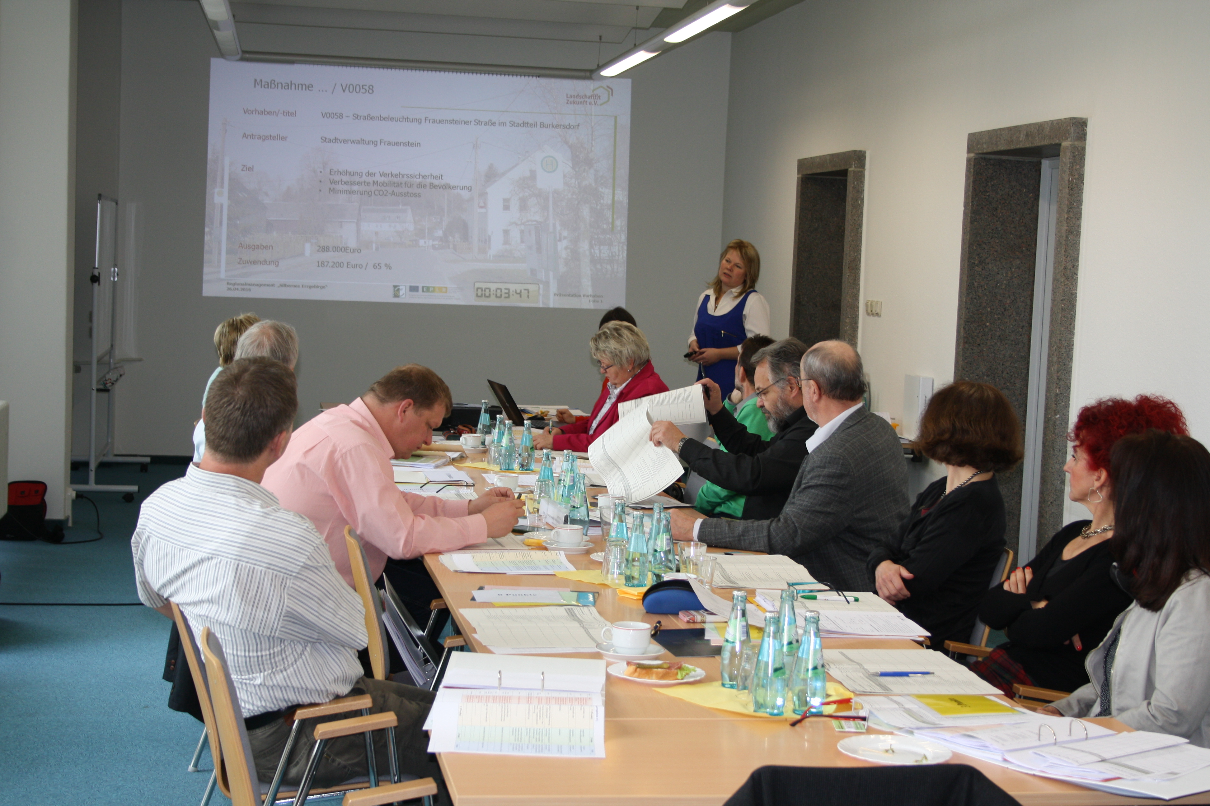 9 Personen sitzen an einem langen Tisch und hören einer Vortragenden zu.