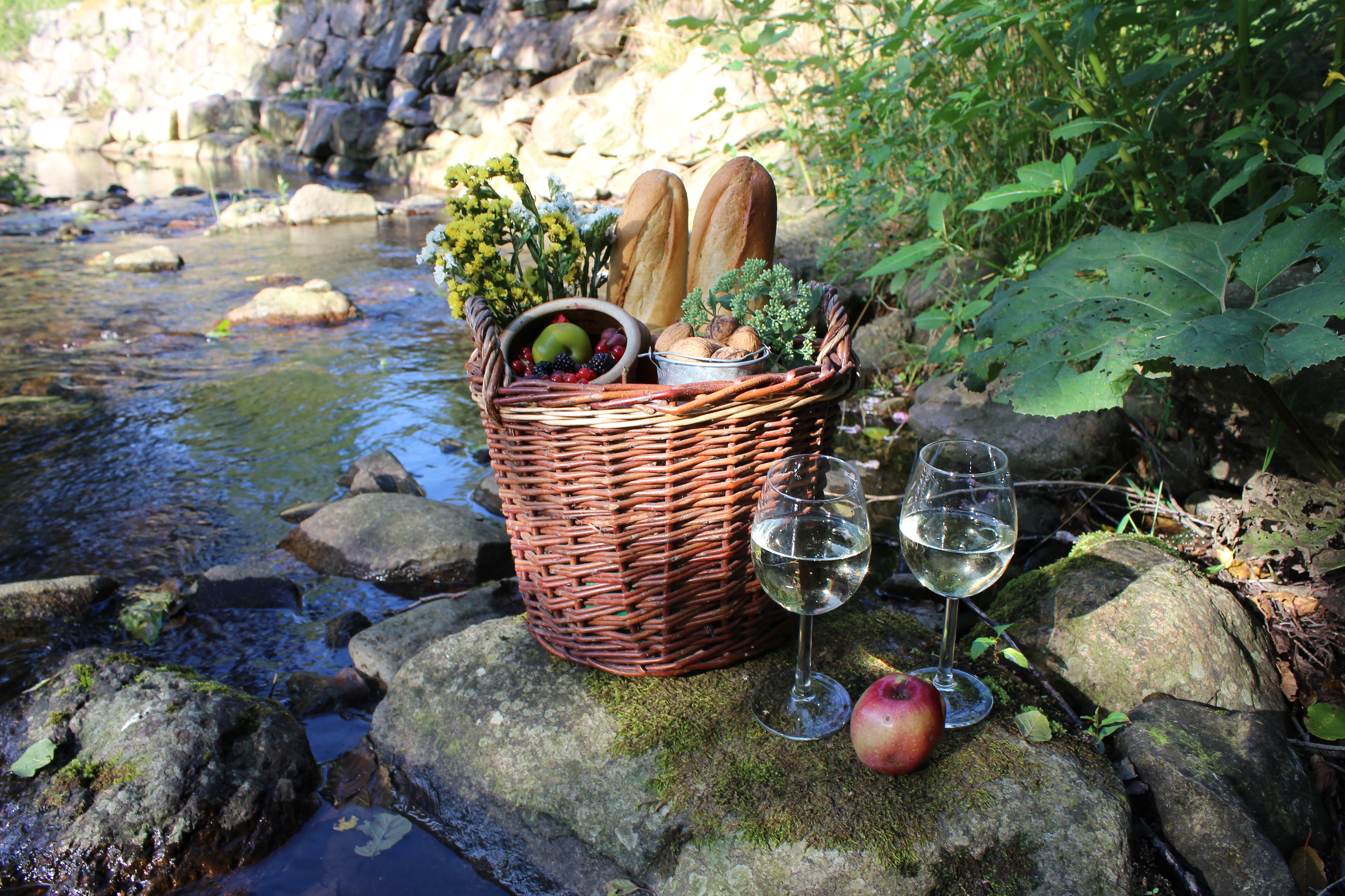 Ein Korb gefüllt mit Baguettes und Obst, daneben zwei Weingläser auf einem Stein an einem Bach.