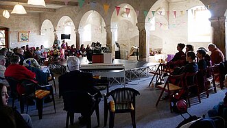 Viele Menschen im einem offenen Raum, in der Mitte eine kleine Bühne.