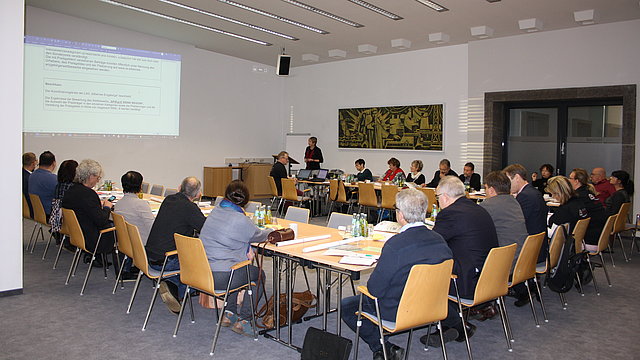 AG Sitzung Großer Raum mit Tischen und Stühlen in U-Form gestellt. An diesem hören Personen einer Vortragenden zu