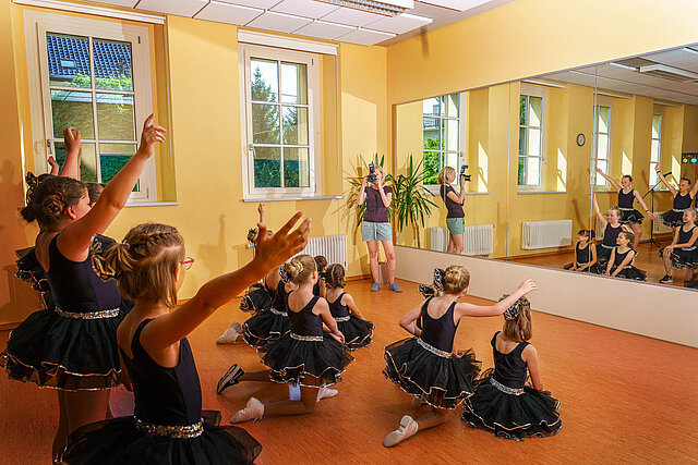 Drei Reihen tanzender Mädchen in schwarzen Tanzkleidern. Die ersten beiden Reihen hocken vor einem Spiegel und die hintere Reihe steht davor.