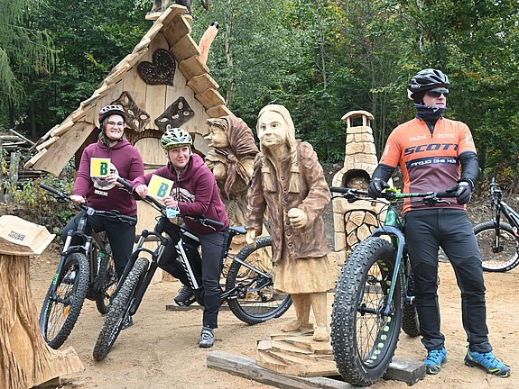 drei Fahrradfahrer stehen um eine lebensgroße und geschnitzte Holzfigur