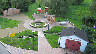 Runder Platz mit Sitzbänken in der Mitte, einem Spielplatz mit Rutsche im Hintergrund und einer Garage.